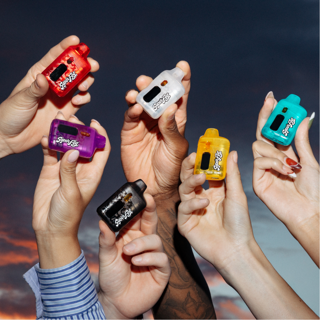 Six small, colorful, compact vape devices held by diverse hands against a sunset sky background.