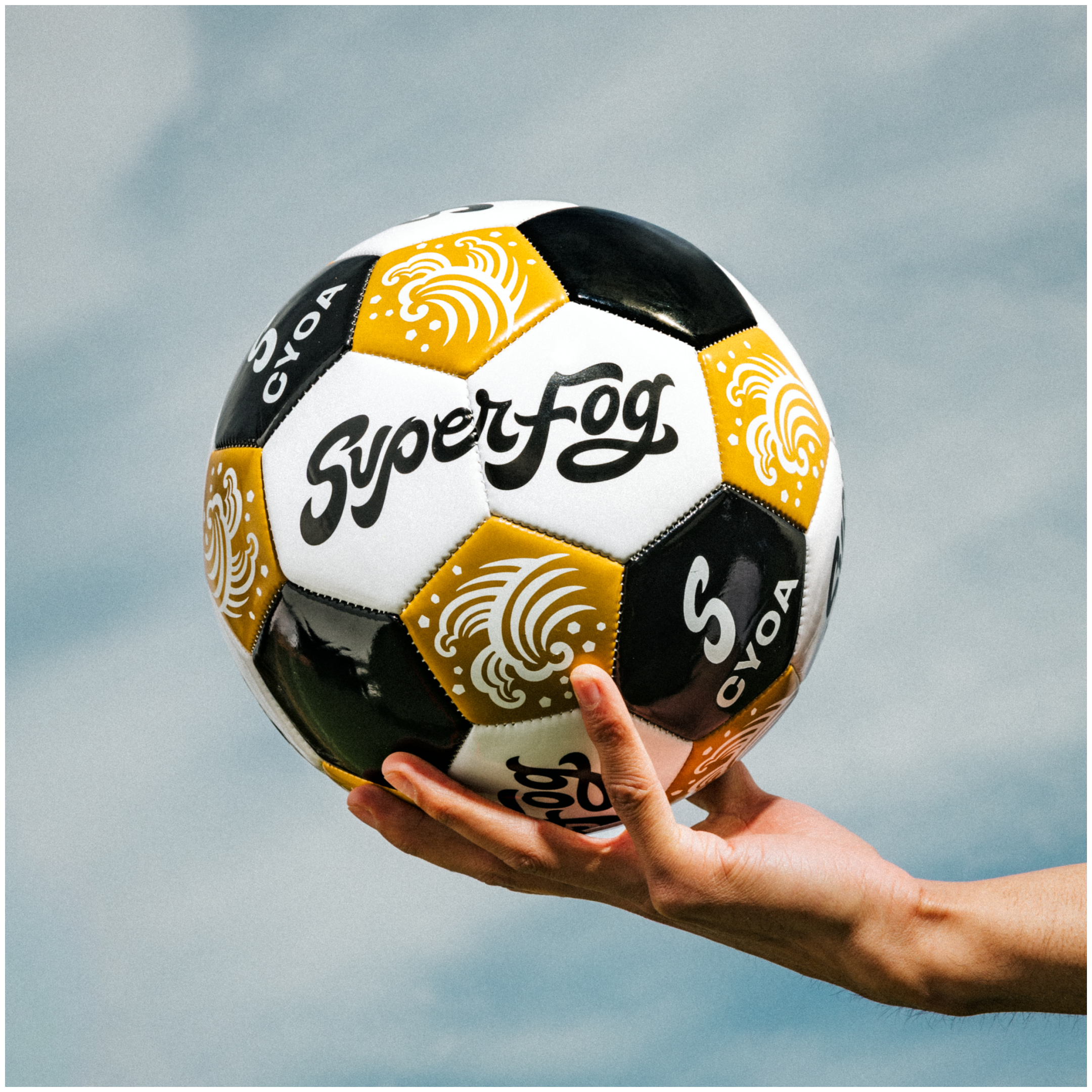 A hand holding a black and white soccer ball with yellow and white cloud-like designs and the word 'SuperFrog' written on it, against a cloudy sky background.