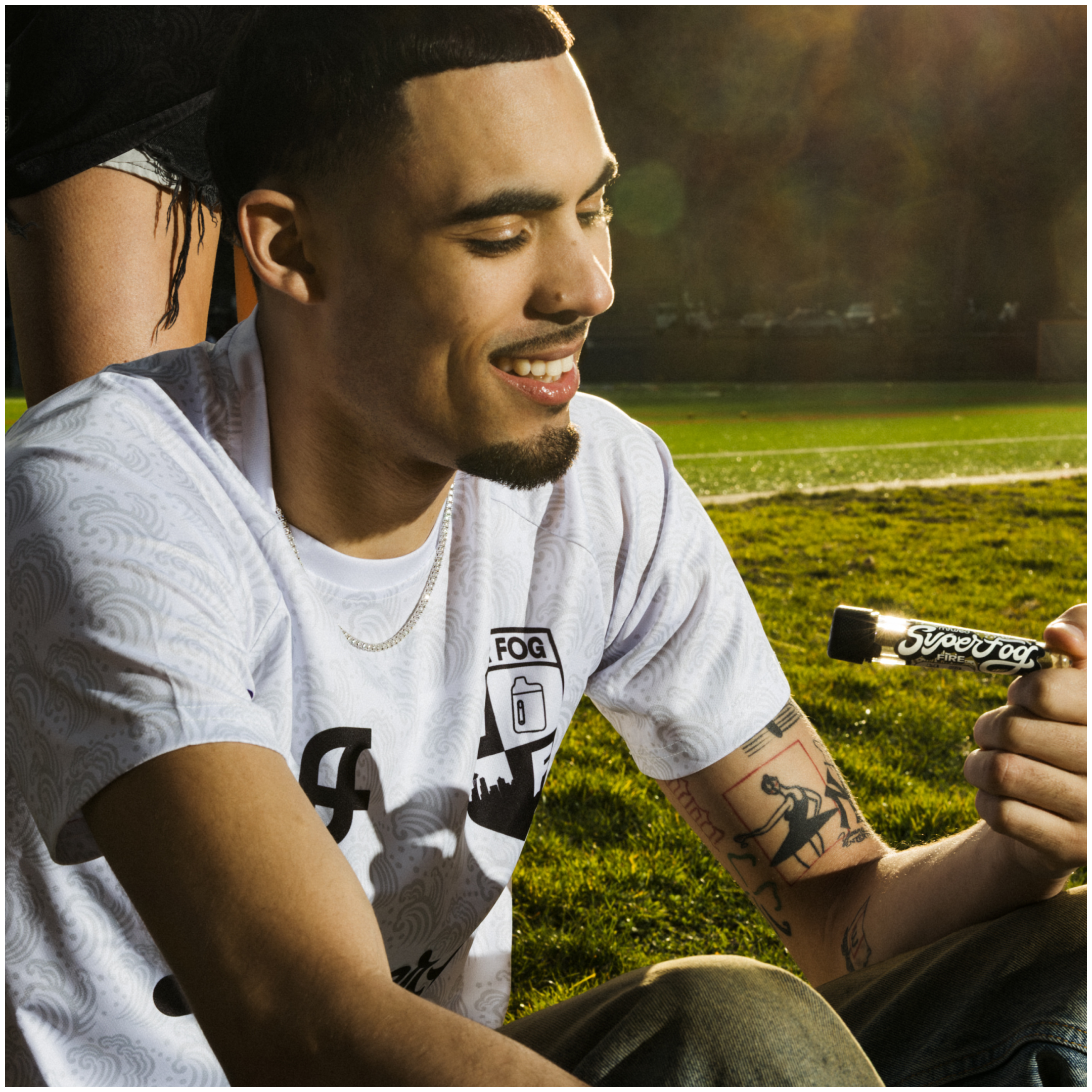 A young man with tattoos on his arms smiling and holding a Super Fog Firework while sitting on grass during sunset.