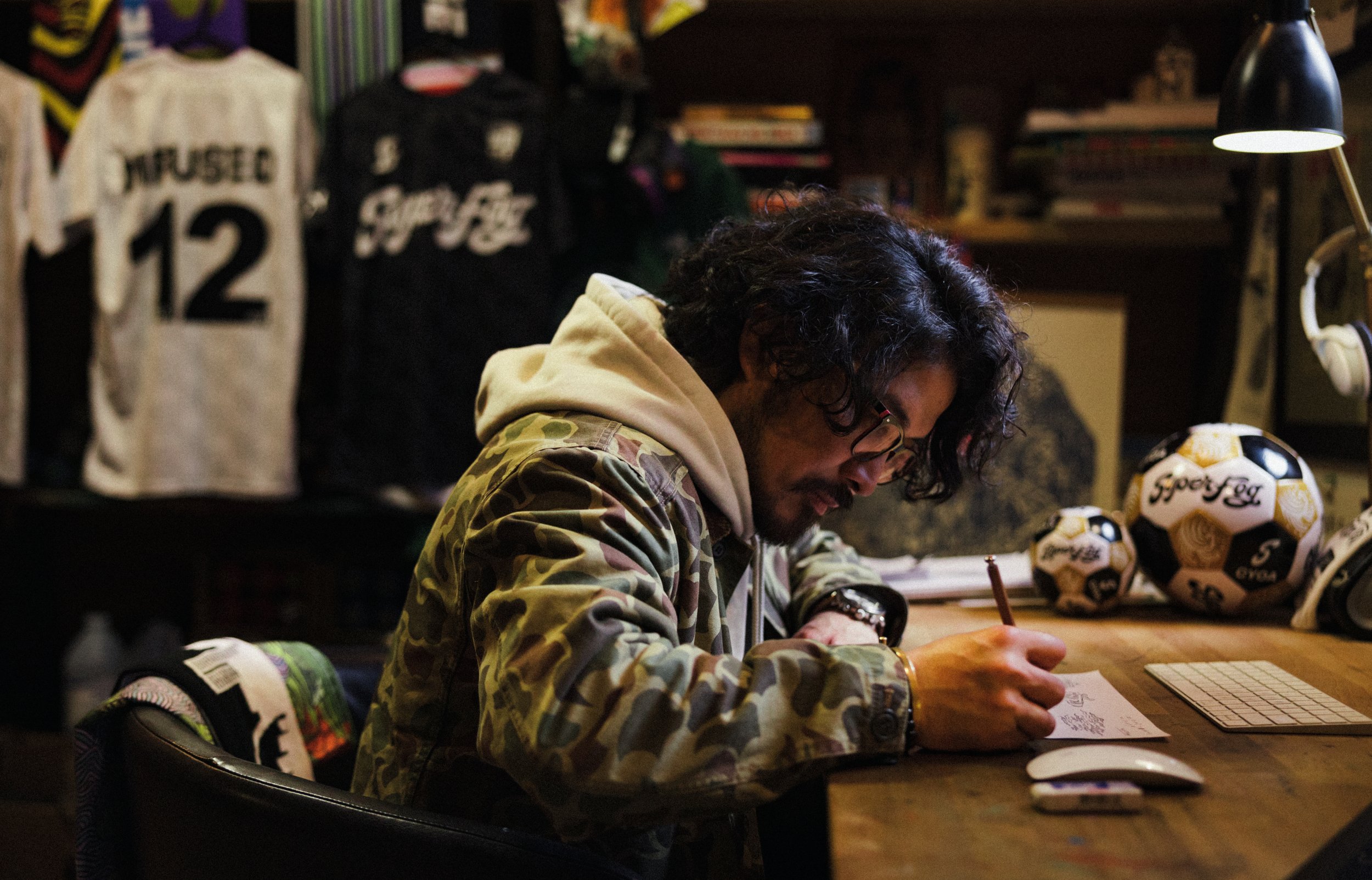 A man with curly dark hair, glasses, and a beard sitting at a wooden desk writing on paper in a cozy room with baseball jerseys and sports memorabilia on shelves behind him. There is a keyboard, a mouse, and a soccer ball on the desk, with a lamp providing light.