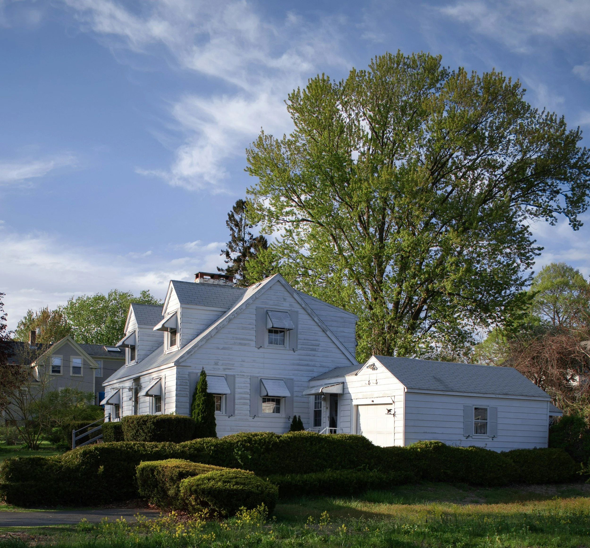 White house with gray roof and shutters, surrounded by green bushes and trees, under a partly cloudy sky.