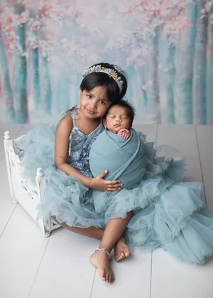 Big sister in a teal tulle dress holding her newborn sibling on a white vintage bed, Bay Area newborn photographer studio session