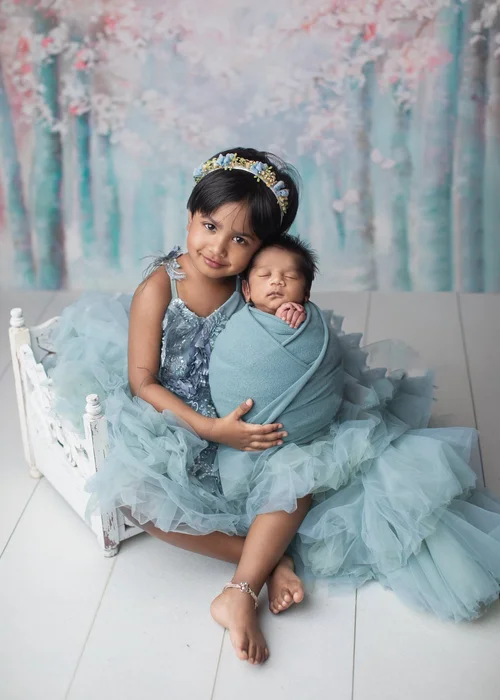 Big sister in a teal tulle dress holding her newborn sibling on a white vintage bed, Bay Area newborn photographer studio session