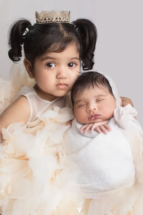 Toddler sister in a crown and cream tulle dress holding her newborn sibling in a bonnet, Mountain View newborn photographer