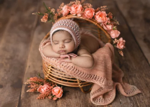 Newborn in a pink knit bonnet resting her chin on her hands in a basket of peach flowers, Mountain View newborn photography studio