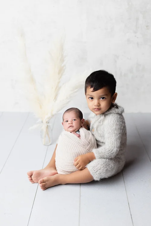 Toddler brother in a grey romper sitting beside his newborn sibling on a white floor, Mountain View newborn photography studio