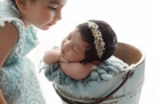 Newborn girl in a bucket prop with her big sister looking down at her on a white backdrop, Mountain View newborn photographer