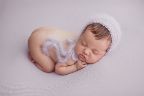 Newborn in a white fluffy bonnet and romper curled up asleep on a lavender backdrop, Bay Area newborn photography session