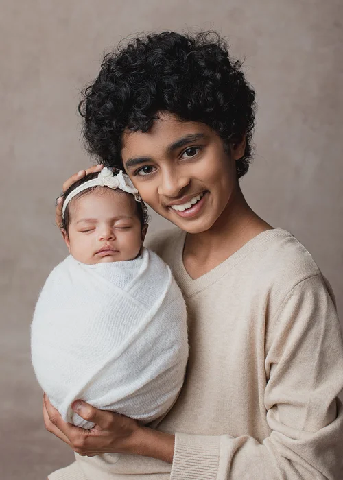 Older sibling in a cream sweater smiling and holding a newborn wrapped in white, Bay Area newborn photography session