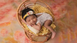 Newborn girl with a yellow headband asleep in a rattan chair on a pink floral backdrop, Bay Area newborn photographer studio session