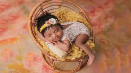 Newborn girl with a yellow headband asleep in a rattan chair on a pink floral backdrop, Bay Area newborn photographer studio session