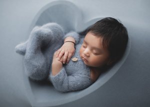 Newborn in a grey-blue knit romper curled up in a grey heart-shaped bowl, Bay Area newborn photographer studio session