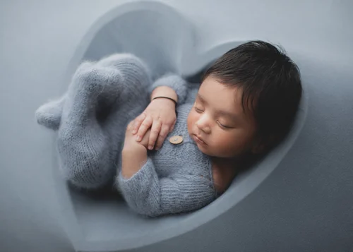 Newborn in a grey-blue knit romper curled up in a grey heart-shaped bowl, Bay Area newborn photographer studio session