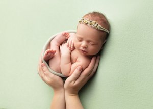 Newborn girl with a beaded headband cradled in a bowl held by parent hands on mint green, Bay Area newborn photographer studio session
