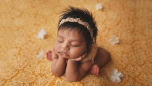 Newborn girl resting her chin on her hands on a golden yellow lace backdrop with small white flowers, Mountain View newborn photography studio
