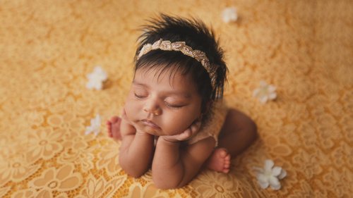 Newborn girl resting her chin on her hands on a golden yellow lace backdrop with small white flowers, Mountain View newborn photography studio