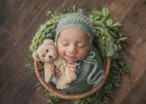 Newborn in a mint knit bonnet hugging a teddy bear in a basket with greenery, Mountain View newborn photographer