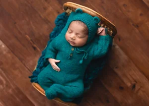 Newborn in a teal bear-hood knit outfit sleeping on a rattan crib bed over dark wood, Bay Area newborn photographer studio session