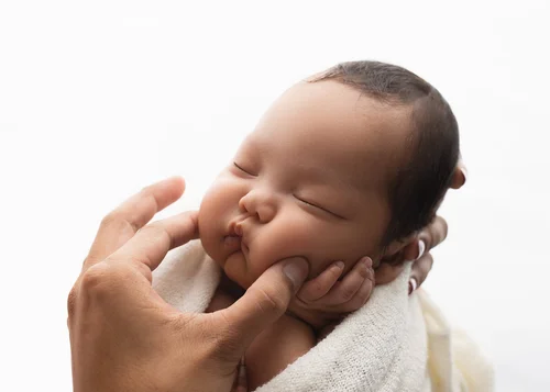 Gentle adult hand cradling the cheek of a sleeping newborn on a white backdrop, Bay Area newborn photography session