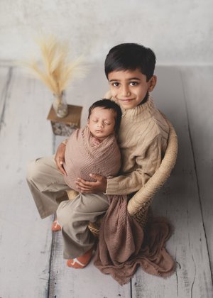 Big brother in a rattan chair holding his swaddled newborn sibling beside pampas grass, Mountain View newborn photography studio