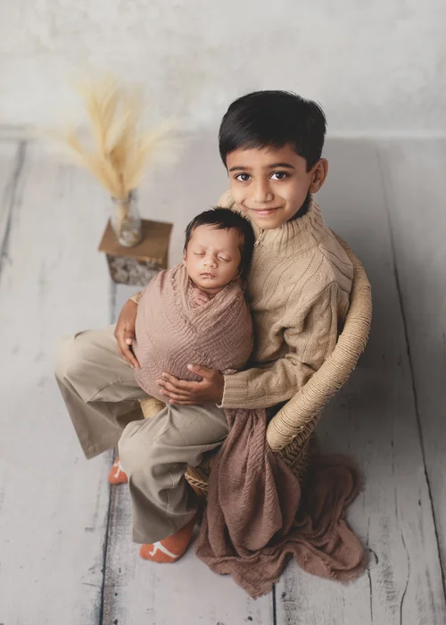 Big brother in a rattan chair holding his swaddled newborn sibling beside pampas grass, Mountain View newborn photography studio