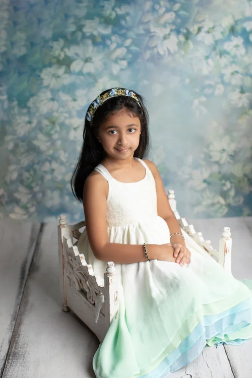 Big sister in a white dress and flower headband sitting on a white vintage bed prop, Mountain View newborn photography studio