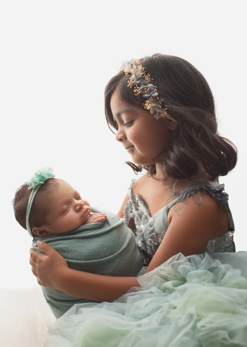 Big sister in a sparkly green dress and floral crown gazing at her sleeping newborn sibling, Mountain View newborn photography studio