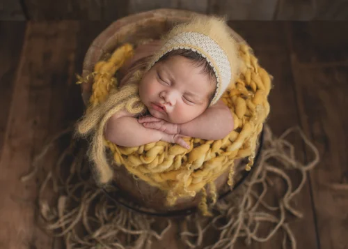 Newborn in a cream knit bonnet nestled in a mustard yellow knit blanket in a basket, Mountain View newborn photography studio