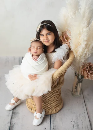 Big sister in a cream tutu dress holding her newborn sibling wrapped in white in a wicker chair, Bay Area newborn photography session