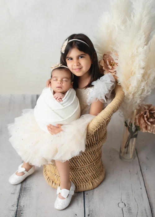 Big sister in a cream tutu dress holding her newborn sibling wrapped in white in a wicker chair, Bay Area newborn photography session