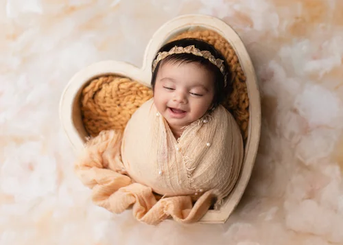 Smiling newborn in a cream wrap in a wooden heart bowl on a peach marble backdrop, Bay Area newborn photographer studio session