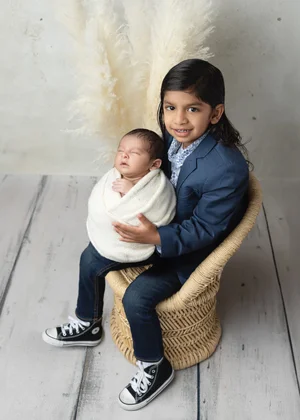 Big brother in a navy suit sitting in a wicker chair holding his newborn sibling in a white wrap, Bay Area newborn photographer studio session