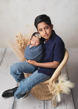 Big brother in a navy shirt and jeans sitting in a wicker chair holding his newborn sibling, Bay Area newborn photography session
