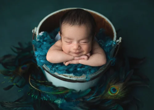 Newborn with hands under his chin in a white bucket with a teal blanket and peacock feathers, Bay Area newborn photographer studio session