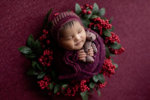 Newborn in a burgundy knit bonnet holding a teddy inside a red berry and greenery wreath, Mountain View newborn photographer