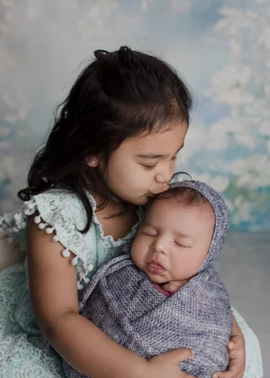 Big sister kissing her newborn sibling swaddled in grey-blue knit, Bay Area newborn photography session