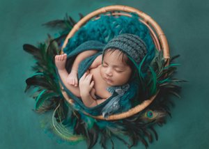 Newborn in a teal knit bonnet inside a feather-lined round basket on a teal backdrop, Bay Area newborn photography session