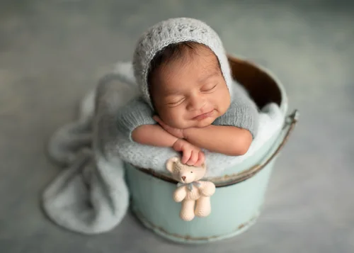 Newborn in a grey-blue knit bonnet in a mint bucket holding a small teddy, Mountain View newborn photography studio