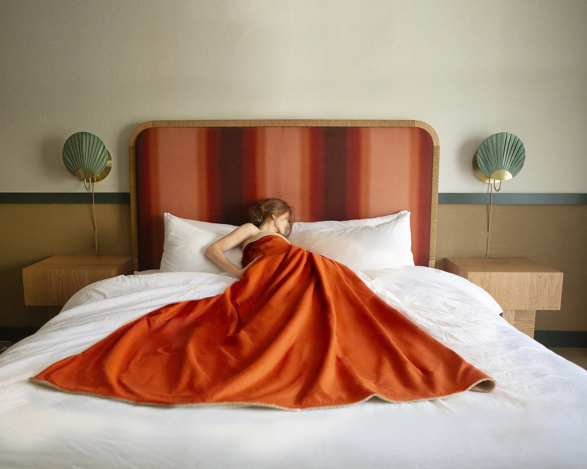 Femme dormant dans un lit avec une couverture orange dans une chambre moderne avec deux lampes vertes et un grand oreiller blanc.