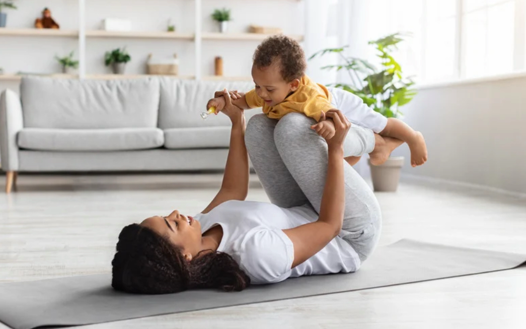 A woman and a young boy playing and laughing on a gray exercise mat in a bright living room with a gray sofa and potted plants.