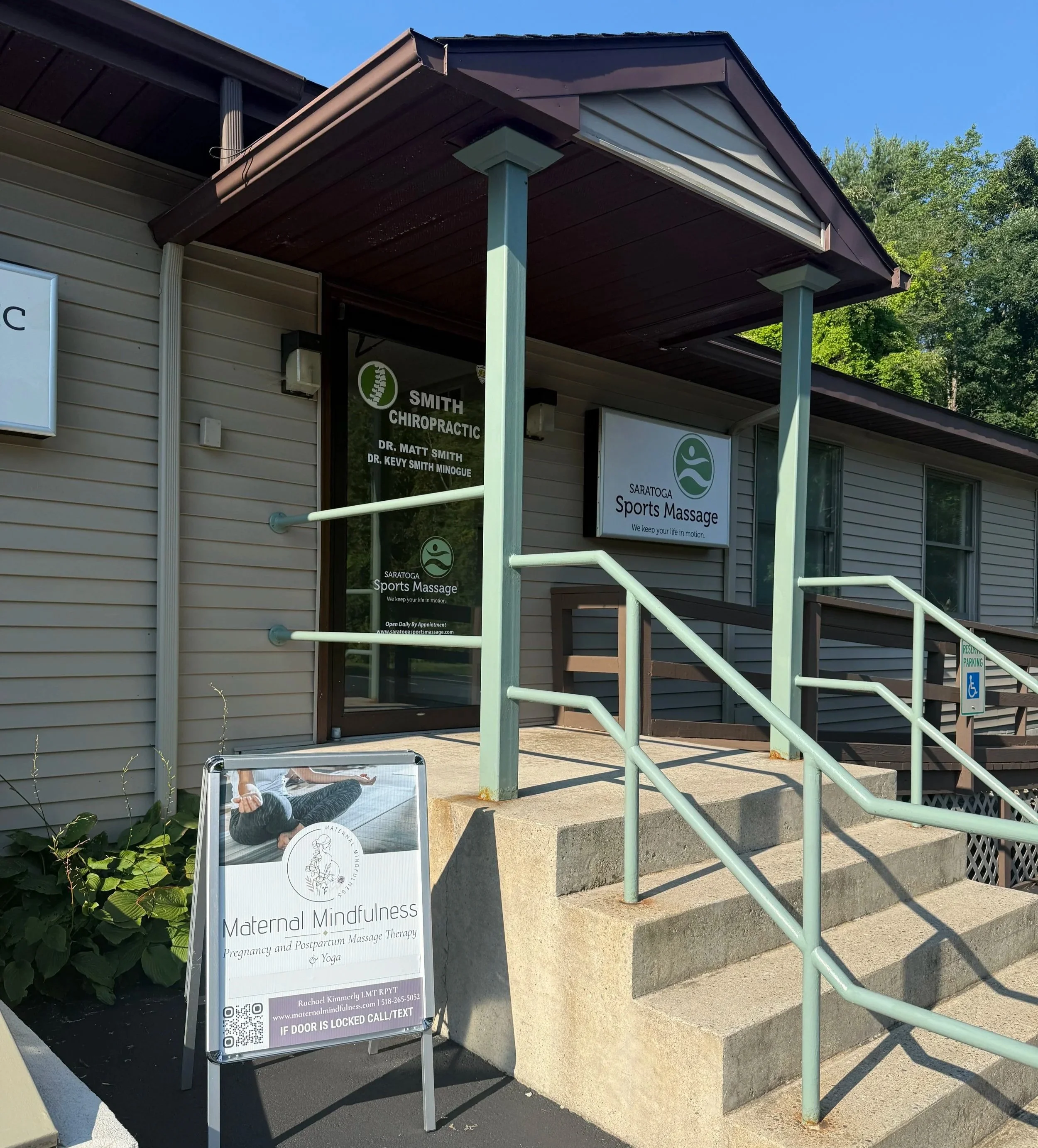 Sign outside a building advertising maternal mindfulness, pregnancy and postpartum massage therapy, and yoga, with the name Rachael Kimmerly and contact information, next to concrete stairs and a handicap accessible parking sign, with green trees and blue sky in the background.