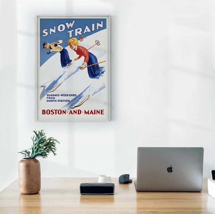 Framed artwork of 2 people skiing with Boston and Maine in large bold red writing, hanging over a computer.