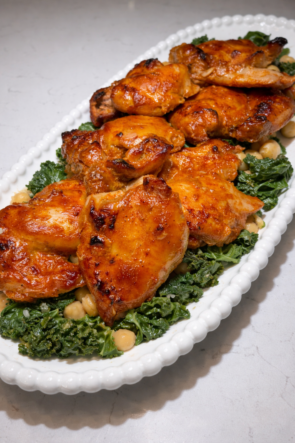 Golden‑brown gochujang‑glazed chicken thighs served on a bed of sautéed kale and chickpeas on a white beaded platter.