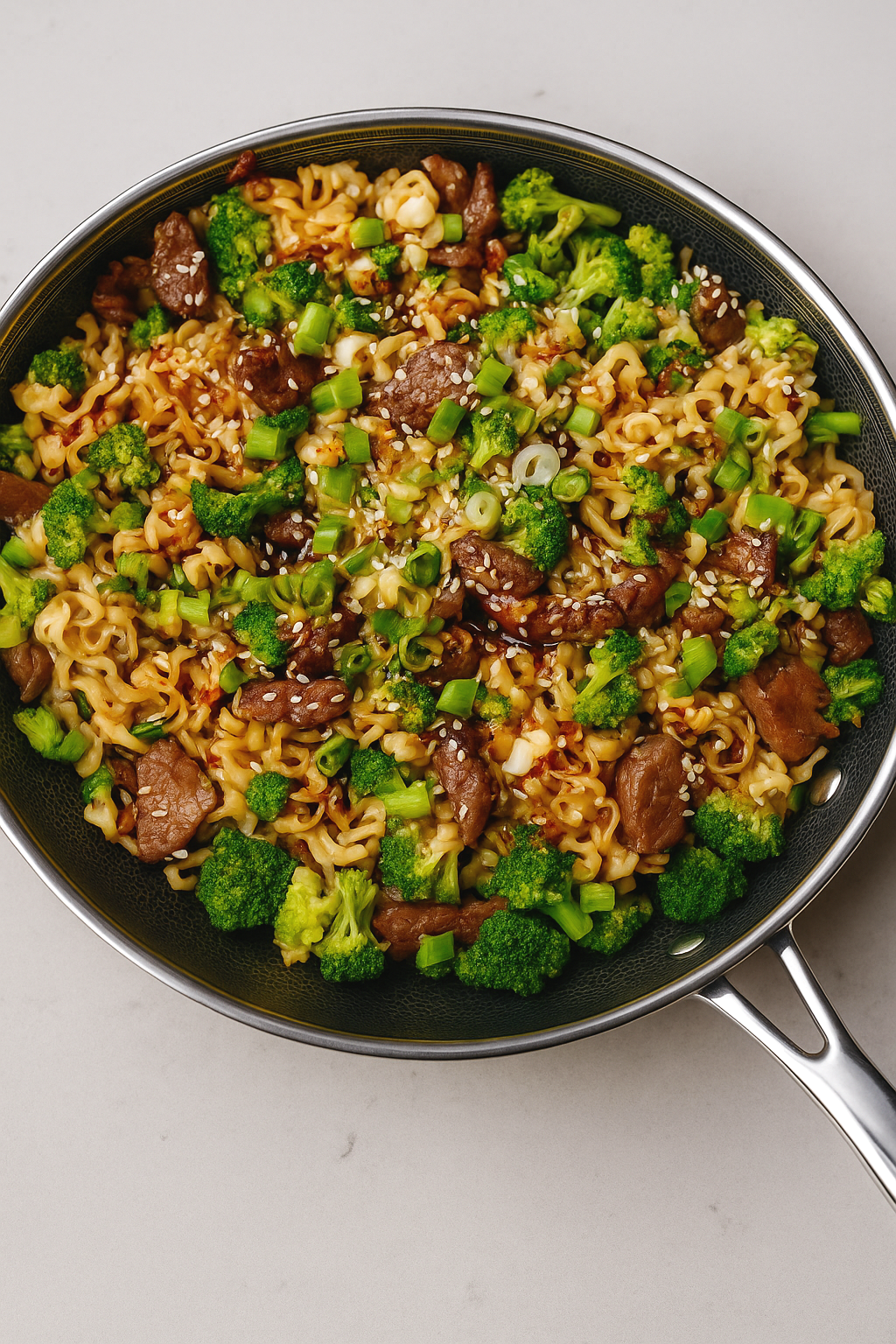 A skillet containing cooked ramen noodles with pieces of beef, broccoli florets, chopped green onions, and sesame seeds sprinkled on top.