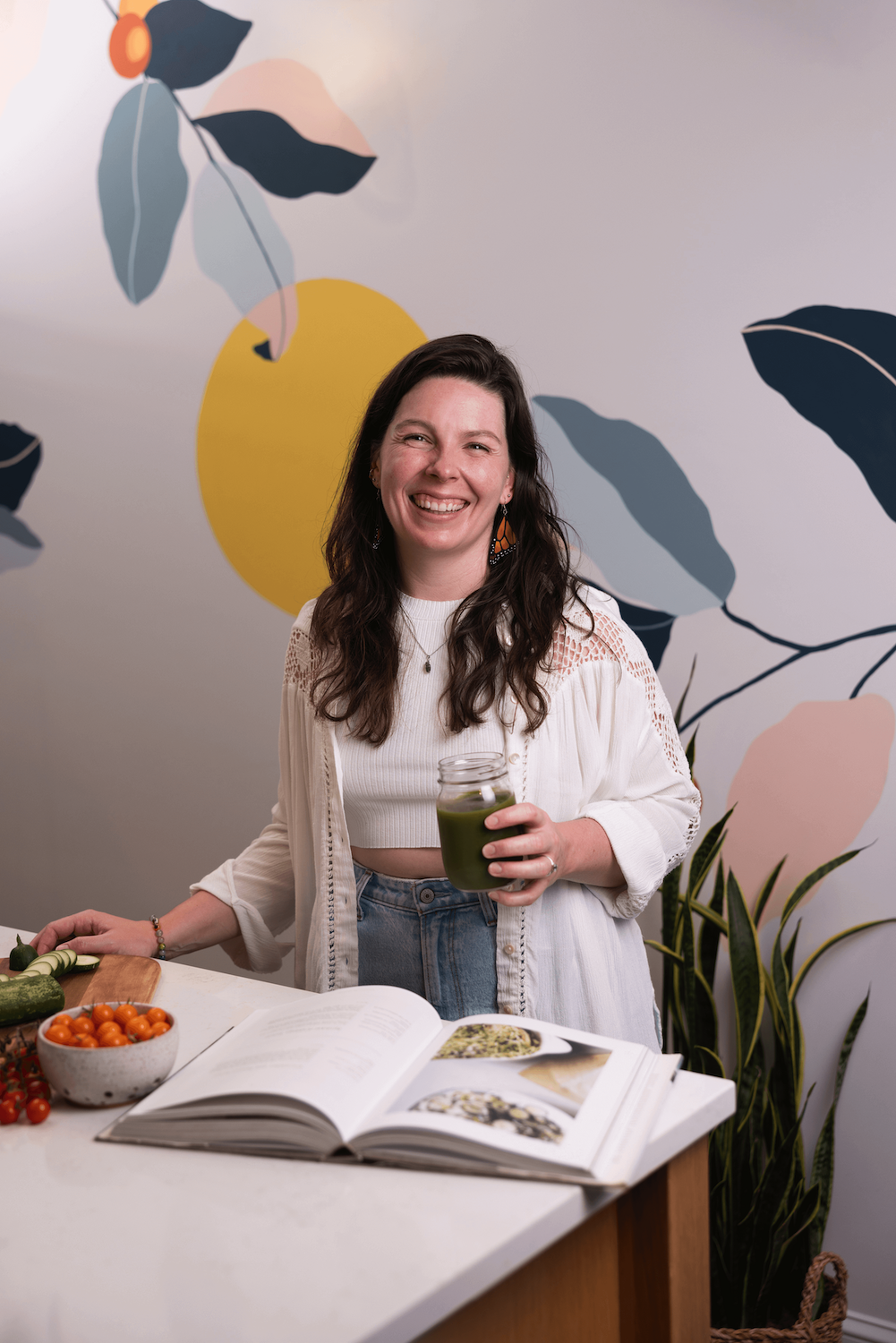 Kaitlyn Carroll, National Board Certified Holistic Health and Wellness Coach offering virtual coaching services, standing in front of colorful mural, holding glass of green juice, smiling at camera