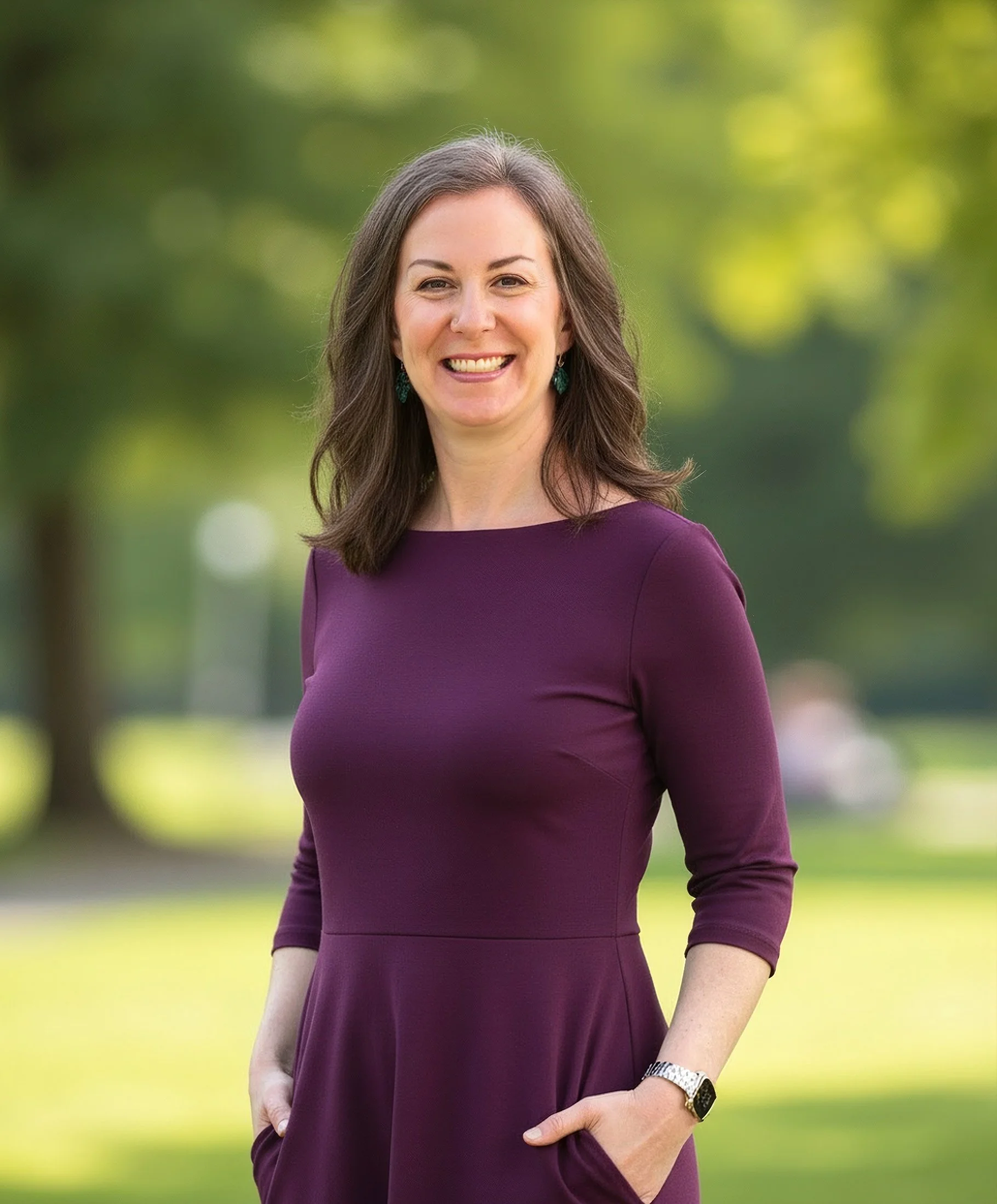 Smiling woman wearing a purple dress standing outdoors with greenery in the background.