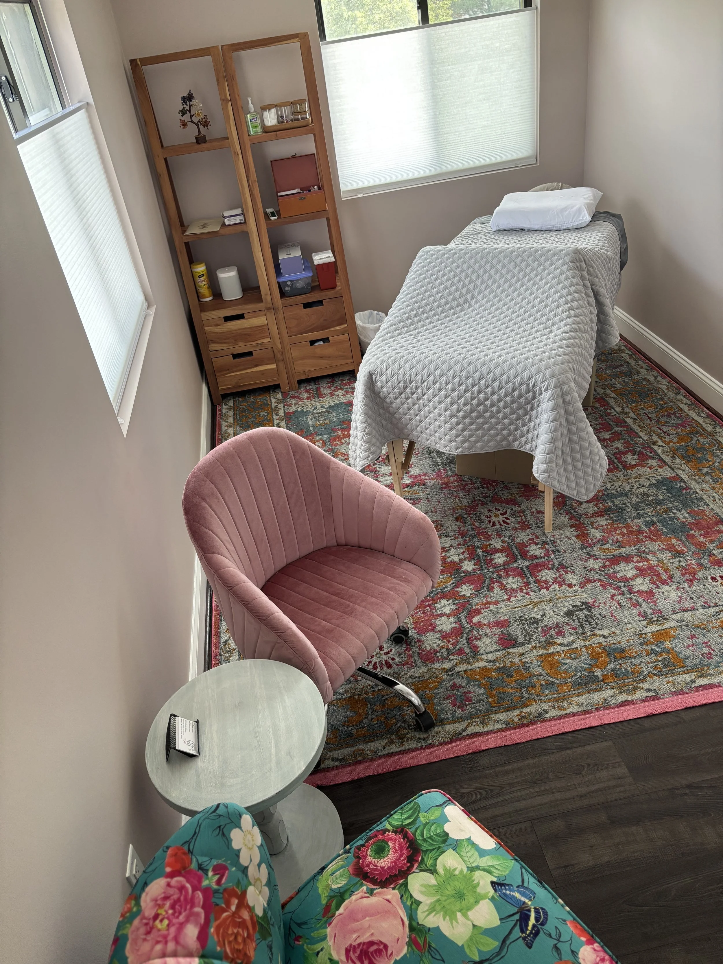Cozy wellness treatment room with a massage table, pink chair, patterned rug, and wooden shelving.