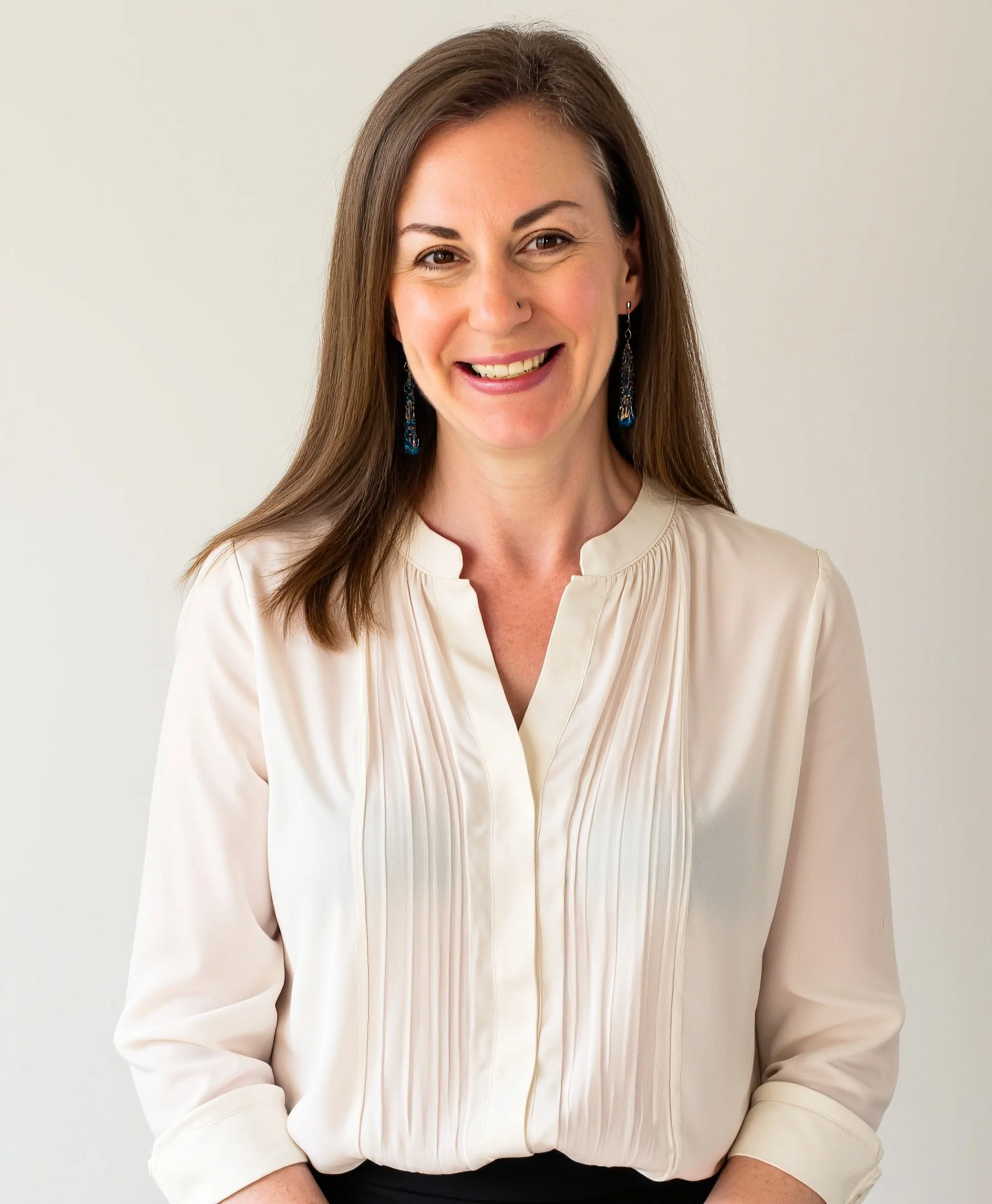 Smiling woman with long brown hair wearing a white blouse against a soft neutral background.