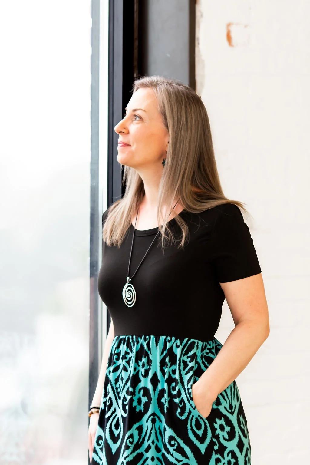 Woman standing by a window wearing a black top and patterned skirt with a pendant necklace.