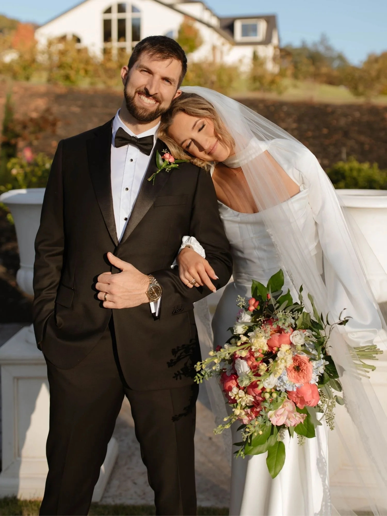 bride and groom with arms linked holding a bouquet of peach, butter yellow, and white